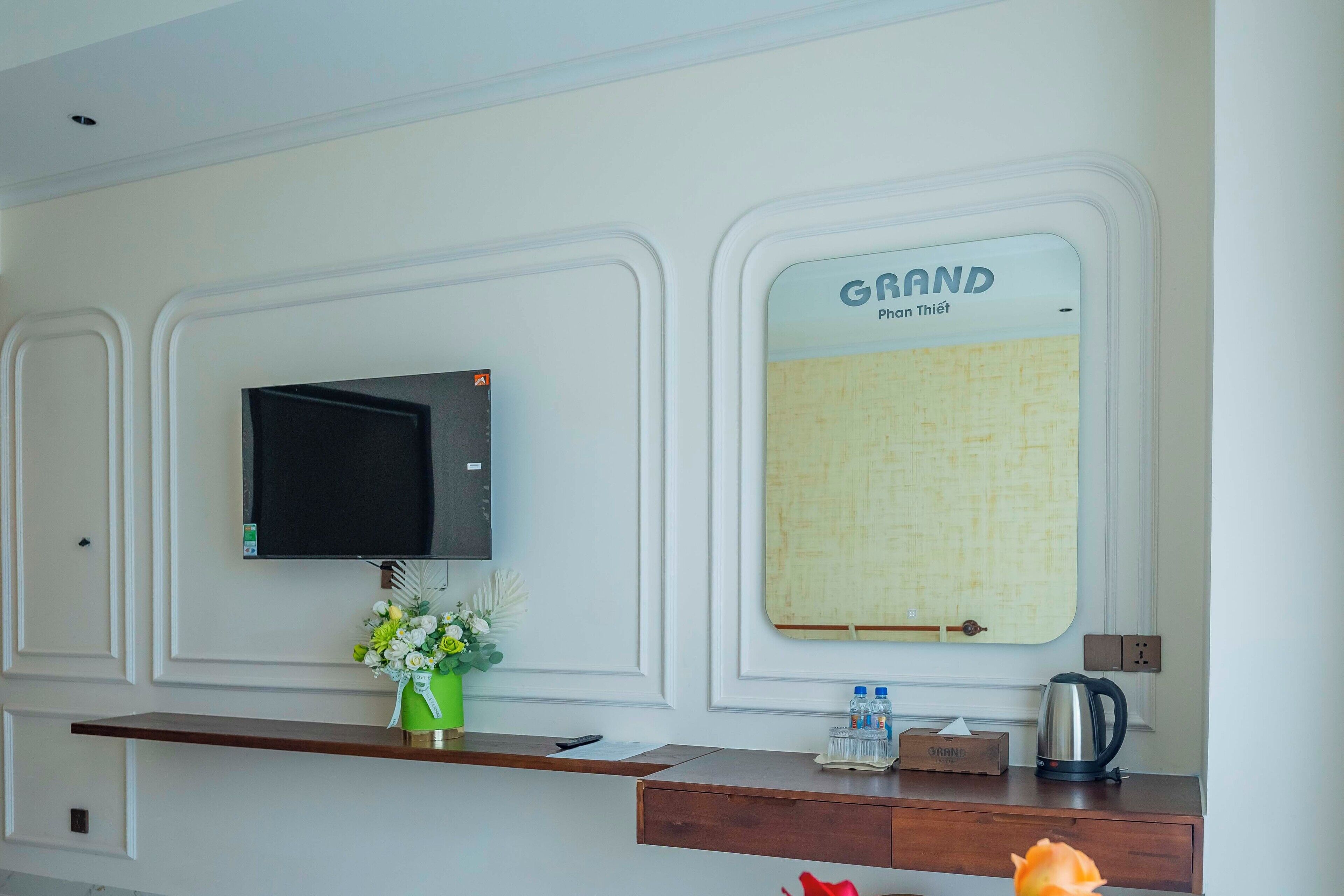 A hotel reception area with a flat-screen TV, a bulletin board, a vase with flowers, a water dispenser, and a coffee maker.