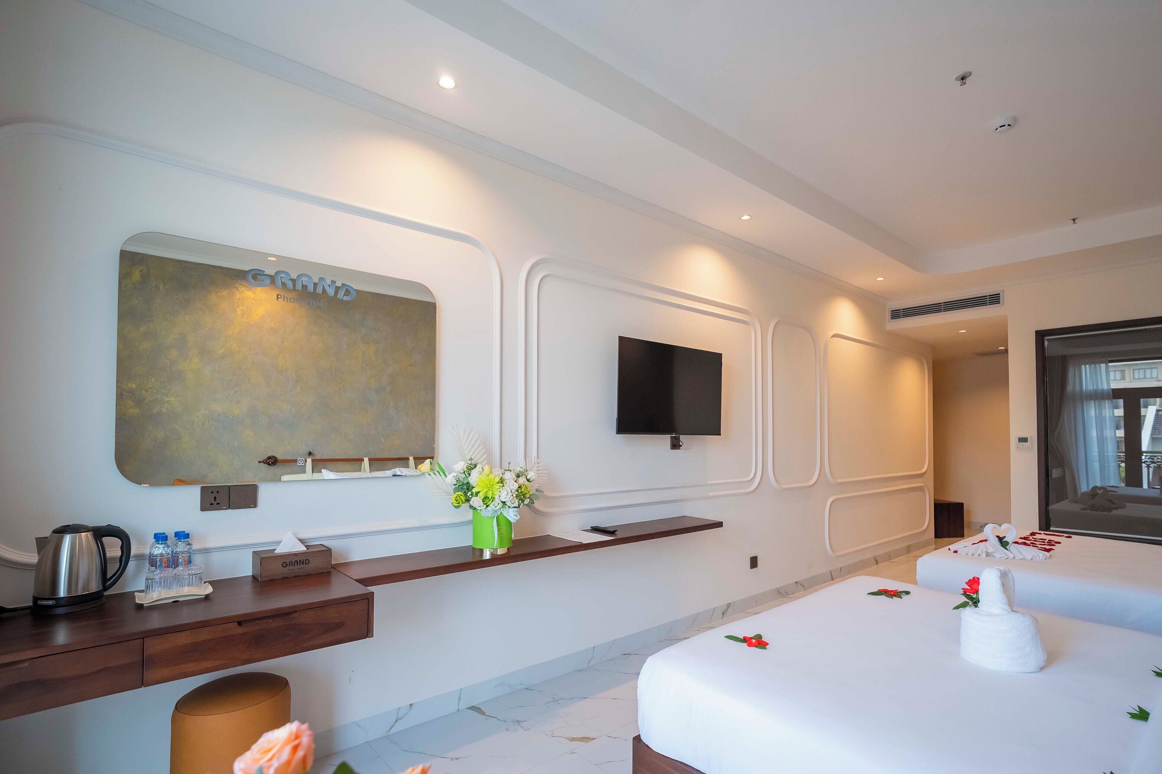 Une chambre d’hôtel moderne avec un grand miroir, un téléviseur à écran plat, un bureau en bois avec une bouilloire, des bouteilles et une boîte de mouchoirs. La chambre est équipée de deux lits avec du linge de maison blanc et d’un vase de fleurs.