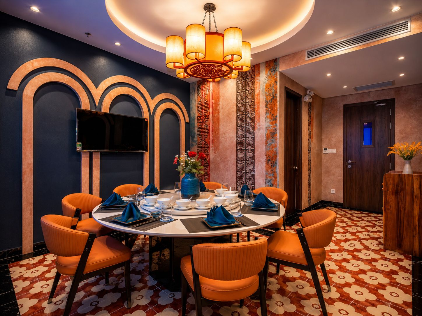 A dining area with a round table, orange chairs, a chandelier, and patterned walls.