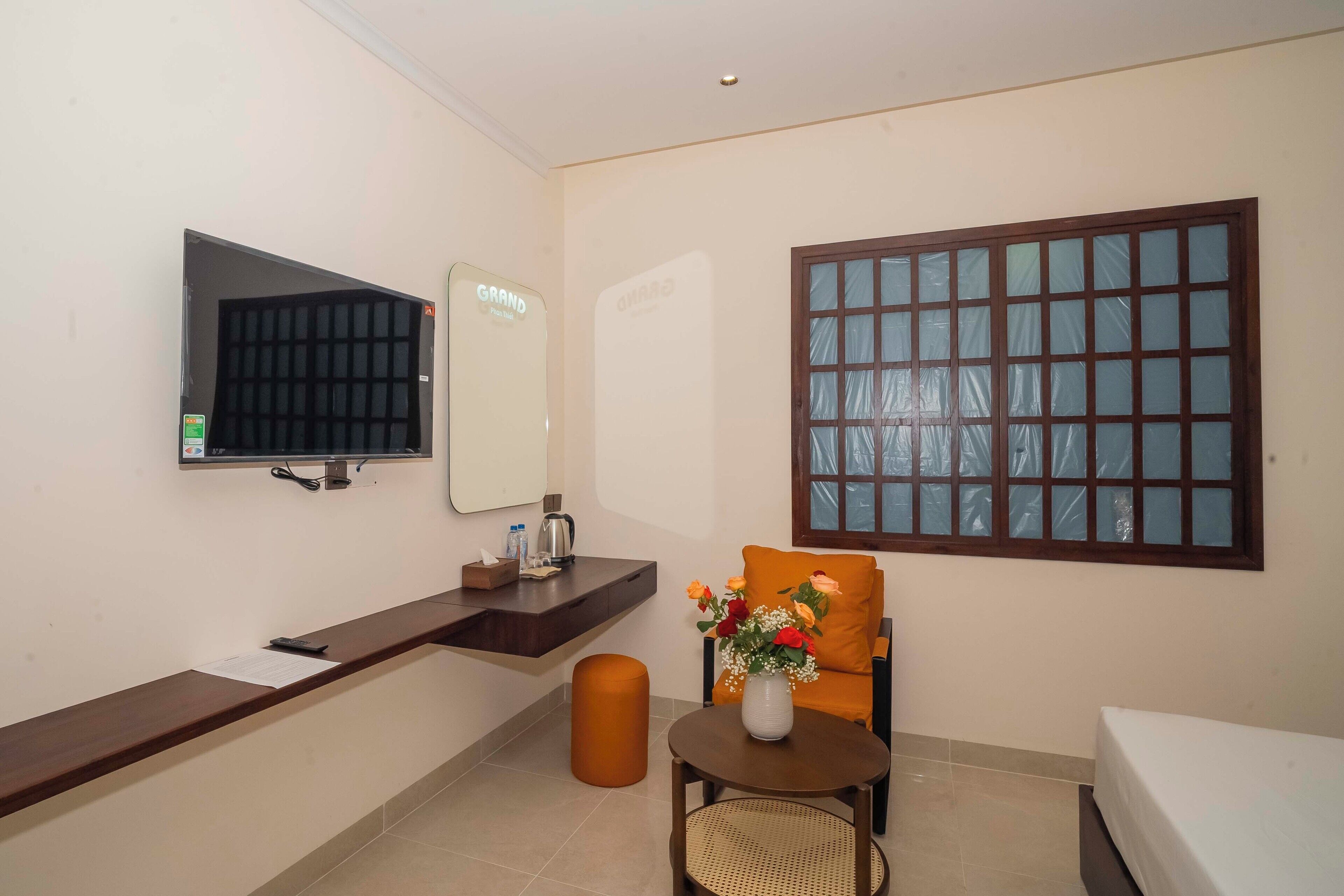 A hotel room with a bed, a small table with a vase of flowers, a chair, a TV, a mirror, a window, and a trash bin.