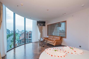 A modern hotel room with a curved floor-to-ceiling window, a leather sofa, a round bed with a white bedspread, and a view of the cityscape.