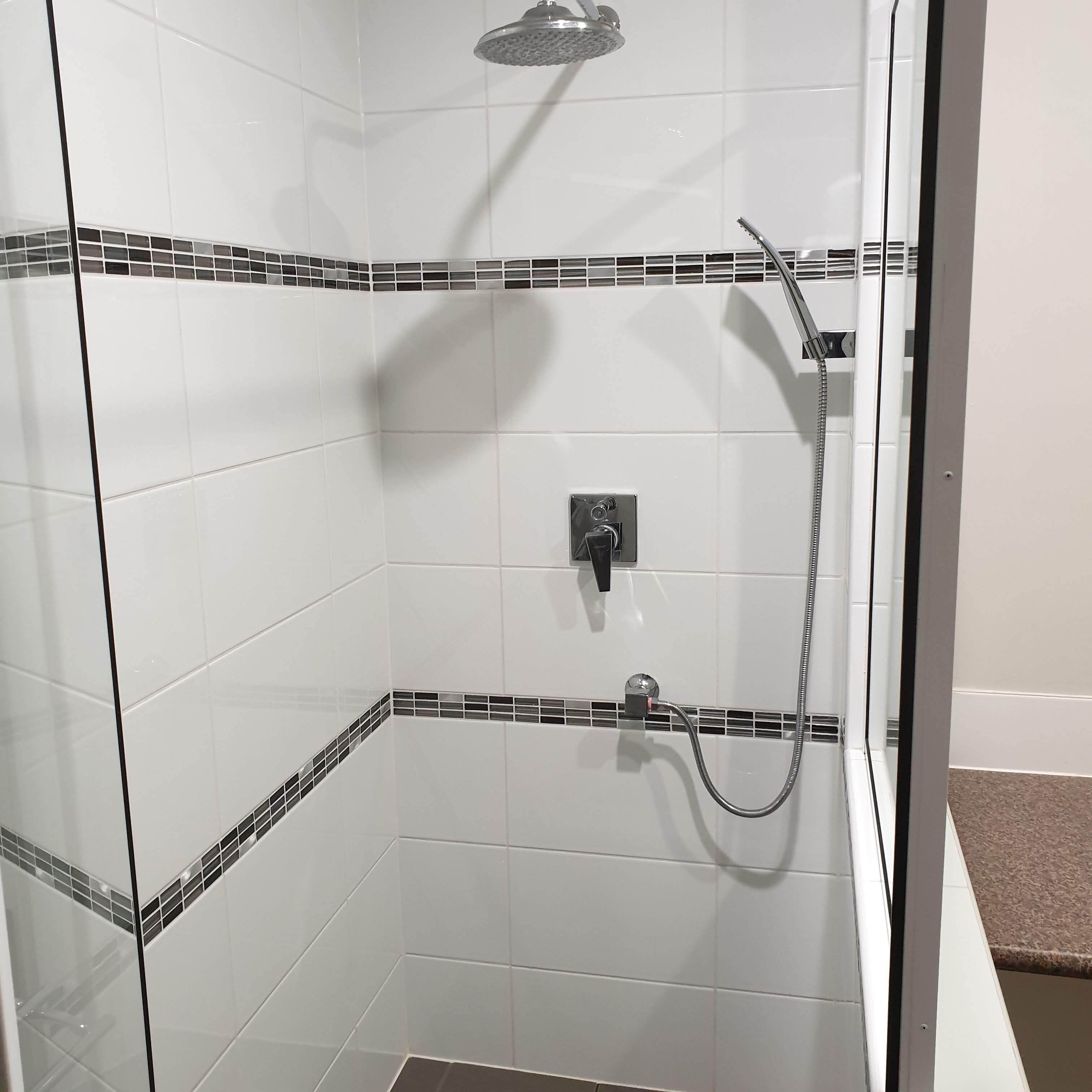 Ducha con cerramiento de vidrio, azulejos blancos y bordes de mosaico negro, cabezal de ducha de mano y grifo empotrado en la pared.