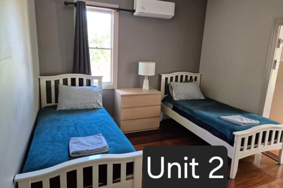 A small, well-lit bedroom with two single beds, a bedside table with a lamp, and a window with curtains.
