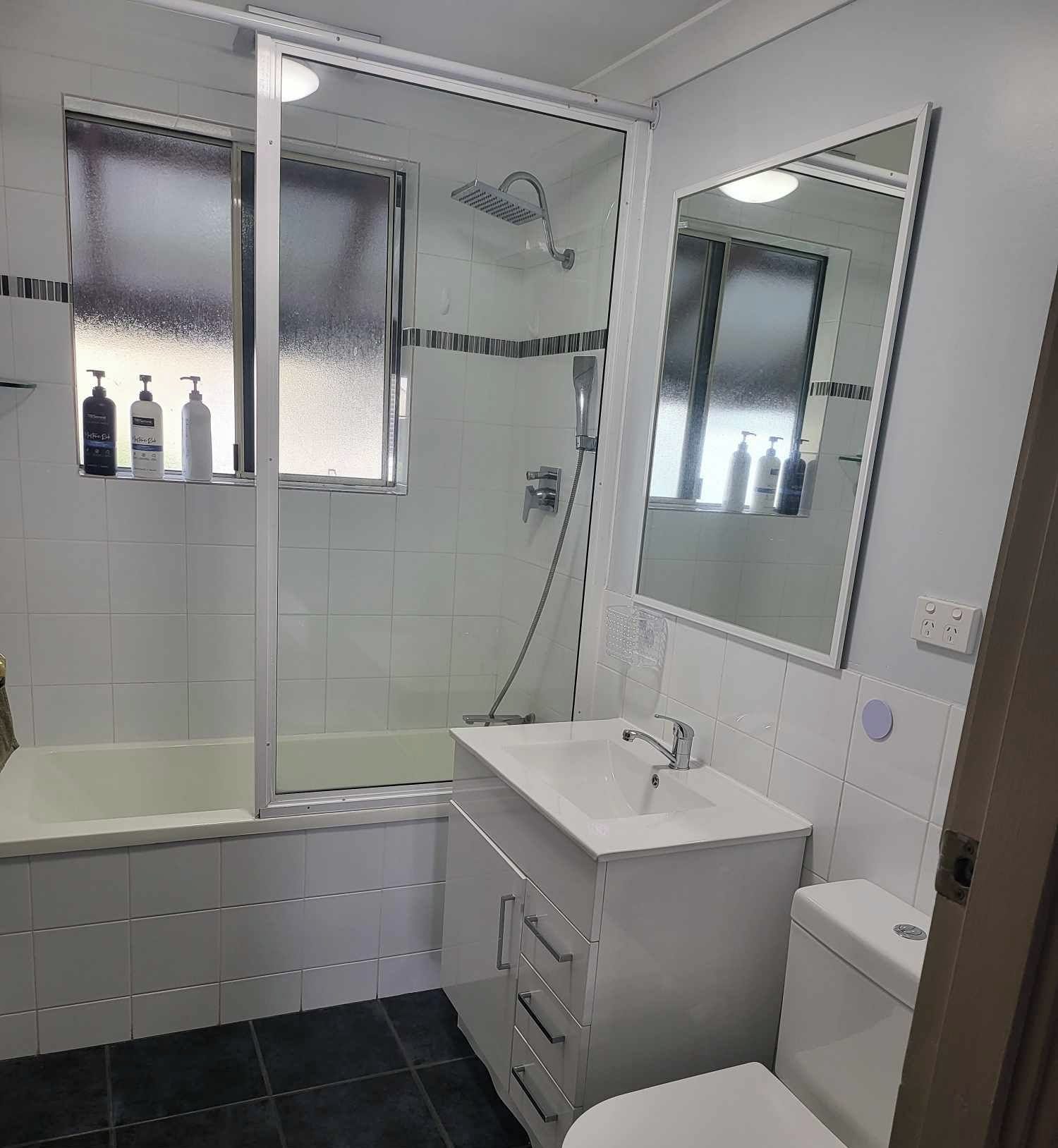 Badeværelse med badekar, håndvask, toilet og bruser. Toiletartikler er anbragt på væggen.