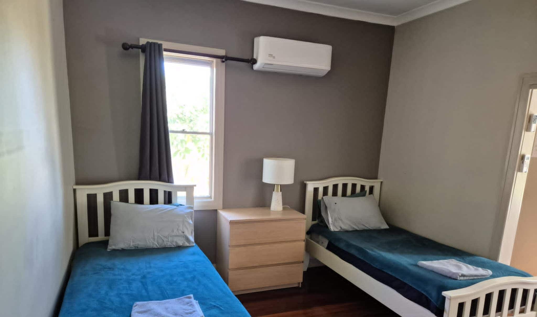 Une petite chambre bien éclairée, avec deux lits simples, une table de chevet avec une lampe et une fenêtre avec des rideaux.