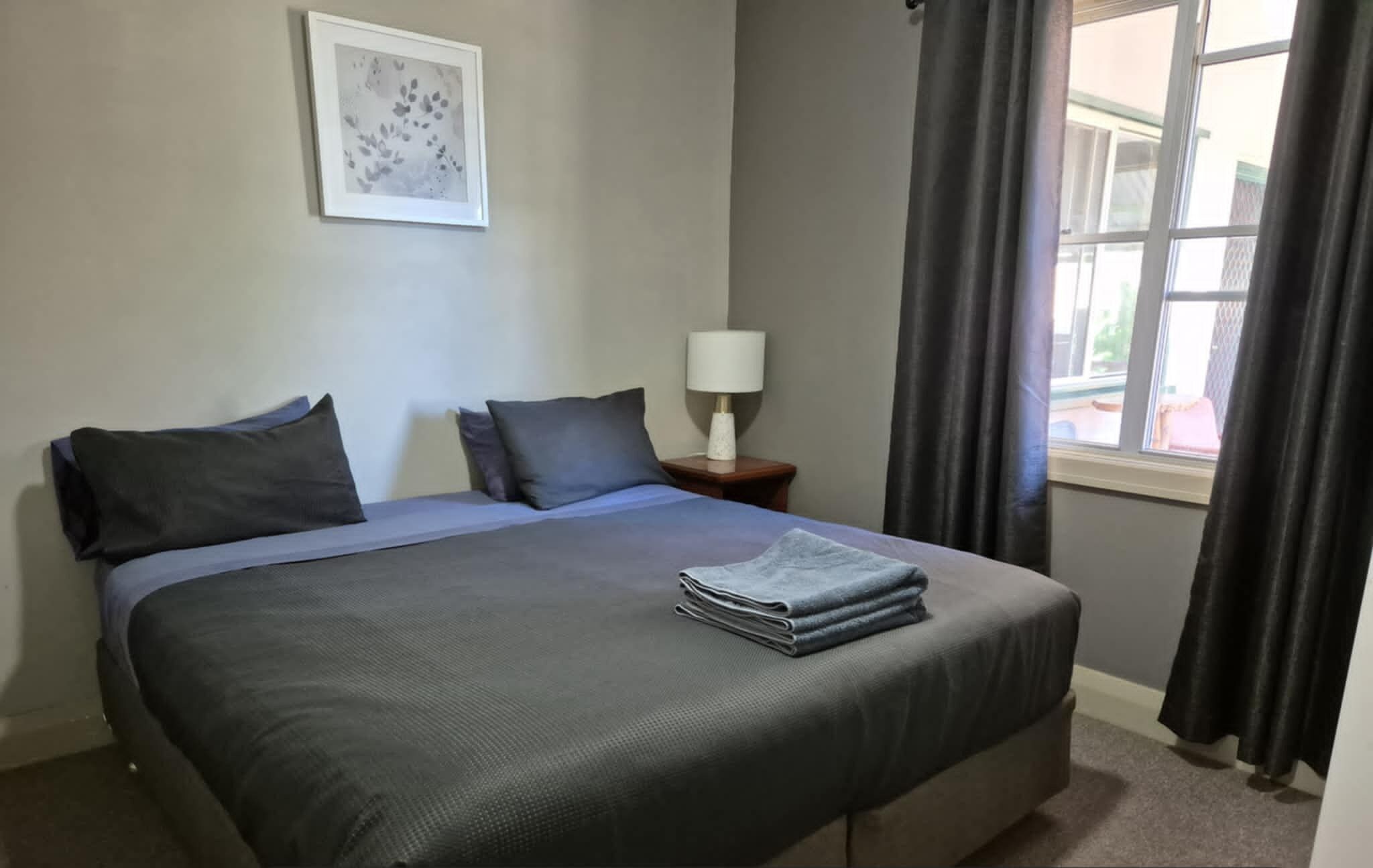 A king-sized bed with a grey bedspread and pillows, a bedside table with a lamp, a framed picture on the wall, and a window with curtains.