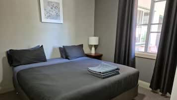 A king-sized bed with a grey bedspread and pillows, a bedside table with a lamp, a framed picture on the wall, and a window with curtains.