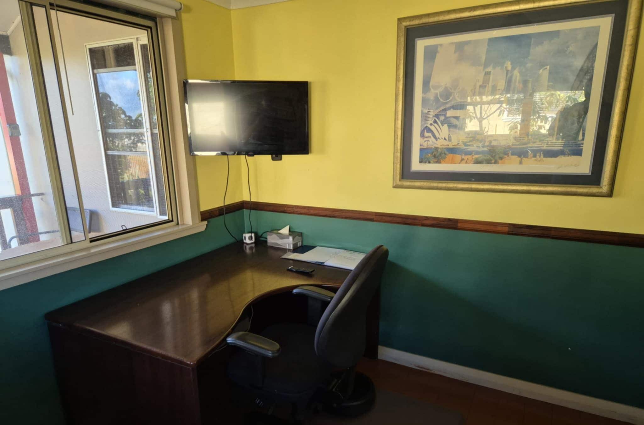 Una habitación con escritorio, silla, ventana y un cuadro en la pared.