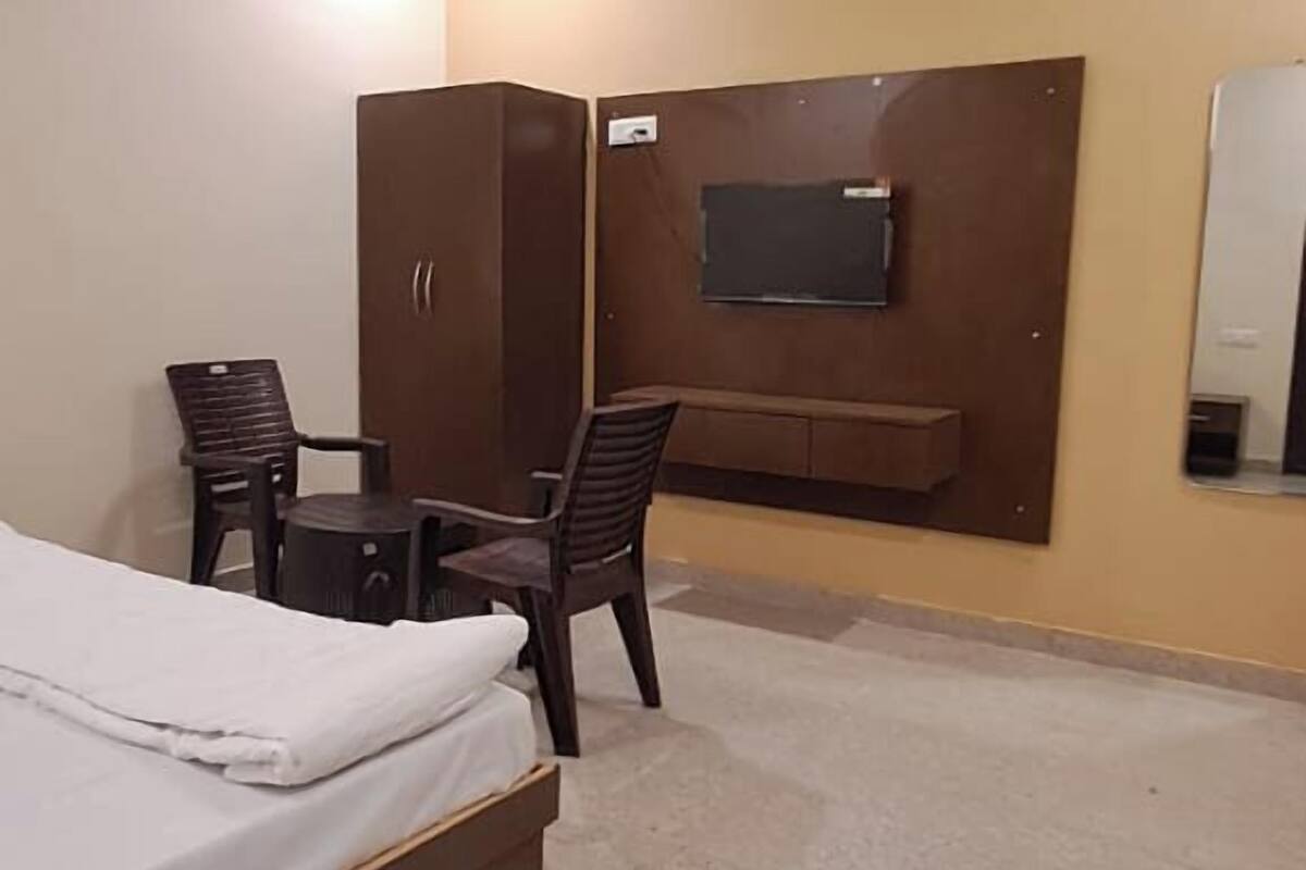 A hotel room with a bed, a desk with two chairs, a wardrobe, a television mounted on the wall, and a ceiling fan.