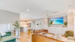 A modern living room with a green sofa, a brown leather sofa, and a green armchair. There is a dining area with a table and chairs, and a kitchen in the background. - New Elevated Home | Pool | 6-Seater Golf Cart (Oak Island)