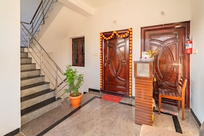 Un hall d’hôtel avec un escalier en marbre, deux portes en bois, un extincteur, une plante en pot et un paillasson.