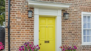 A brick building with a yellow door, two potted flowers, and a window.