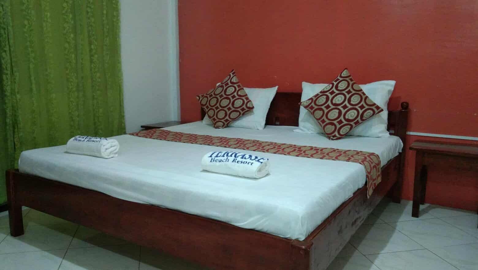 Una cama bien tendida con una colcha blanca y dos almohadas estampadas, cabecera de madera y una cortina verde a la izquierda.