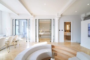 Interior moderno de hotel com área de refeições, sala de estar com sofá curvo e um quarto visível por uma porta de vidro.