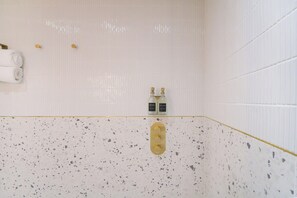 A bathroom with white tiled walls, a speckled floor, two wall-mounted faucets, and a towel rack with towels. - Blanco | The Heim (Greater Manchester)