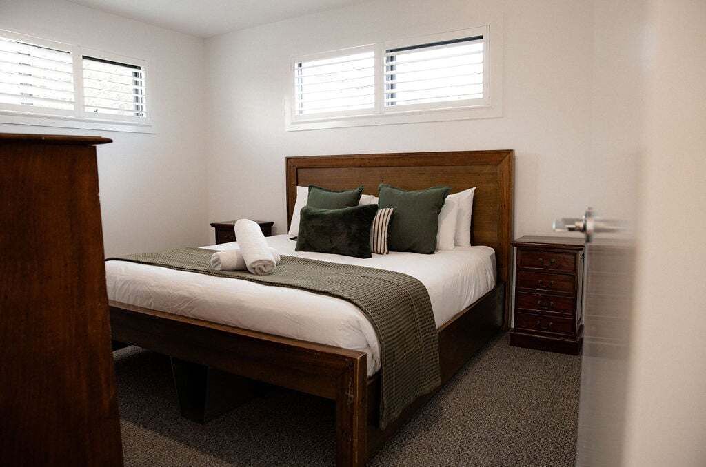 Une chambre à coucher avec un lit en bois, une literie blanche, des coussins verts, une commode en bois et deux fenêtres équipées de stores.