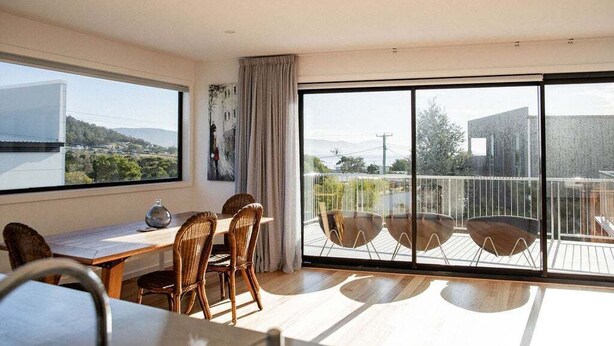 Amplia habitación con un gran ventanal corredero que da a un balcón, un comedor con sillas de mimbre y vistas del paisaje circundante.