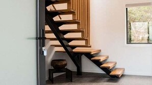 Una escalera moderna con peldaños de madera y barandillas negras, que conduce a una habitación con un gran ventanal y una pequeña mesa.