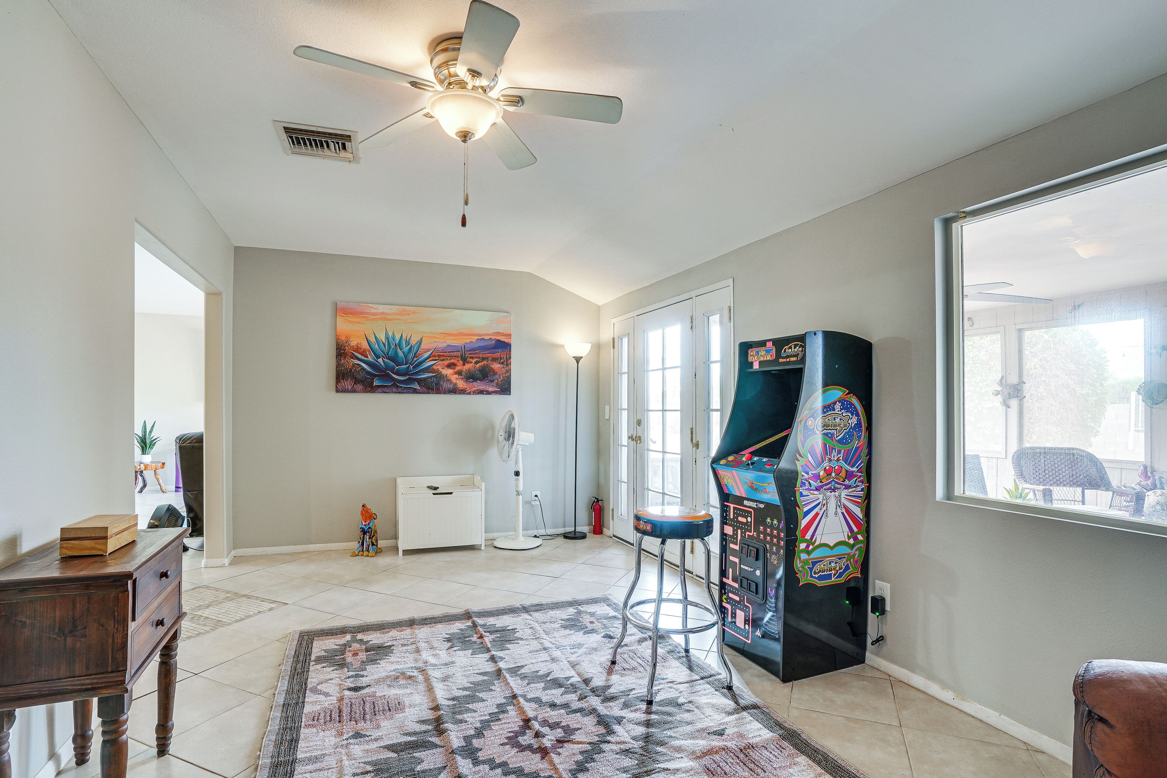 Une pièce spacieuse avec un ventilateur de plafond, une grande fenêtre, une borne d’arcade aux couleurs vives et un tableau accroché au mur.