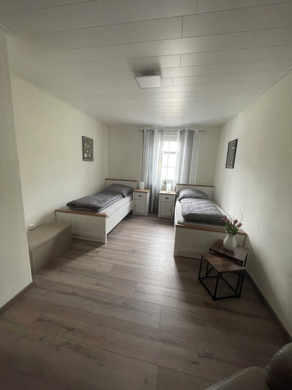 Una habitación pequeña y bien iluminada con dos camas individuales, una mesita con un florero y una ventana con cortinas.