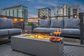Une terrasse sur le toit avec un foyer en verre, des canapés gris et une table en verre sur laquelle sont disposés des fruits et une boisson.