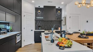 A modern kitchen with dark cabinetry, a central island with fruit, and a dining area with a wooden table. - Music Row - Luxury Home w/ 5 en-suites! (Nashville)