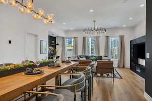 A modern dining area with a long wooden table, cushioned chairs, and pendant lighting.
