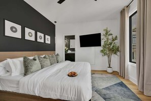 A modern bedroom with a large bed, a flat-screen TV, a potted plant, and a view of the city.