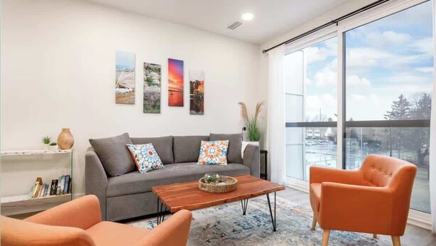 A modern living room with a grey sofa, orange armchairs, and a wooden coffee table. There are three framed pictures on the wall and a large window offering a view of the city.