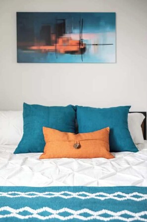 Un lit avec deux oreillers turquoise et un oreiller orange, une couverture à motifs bleus et blancs, et un tableau représentant un bateau accroché au mur.