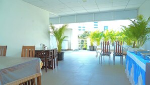 A spacious indoor area with a long table covered with a blue tablecloth, surrounded by wooden chairs. There are potted plants and a glass partition in the background. - Sweet Crystal Homes C V Raman Nagar (Bengaluru)