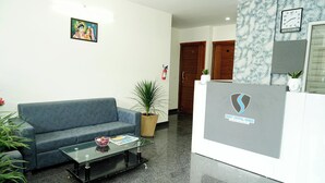 A reception area with a blue sofa, a glass coffee table, a potted plant, and a reception desk with a logo. - Sweet Crystal Homes C V Raman Nagar (Bengaluru)
