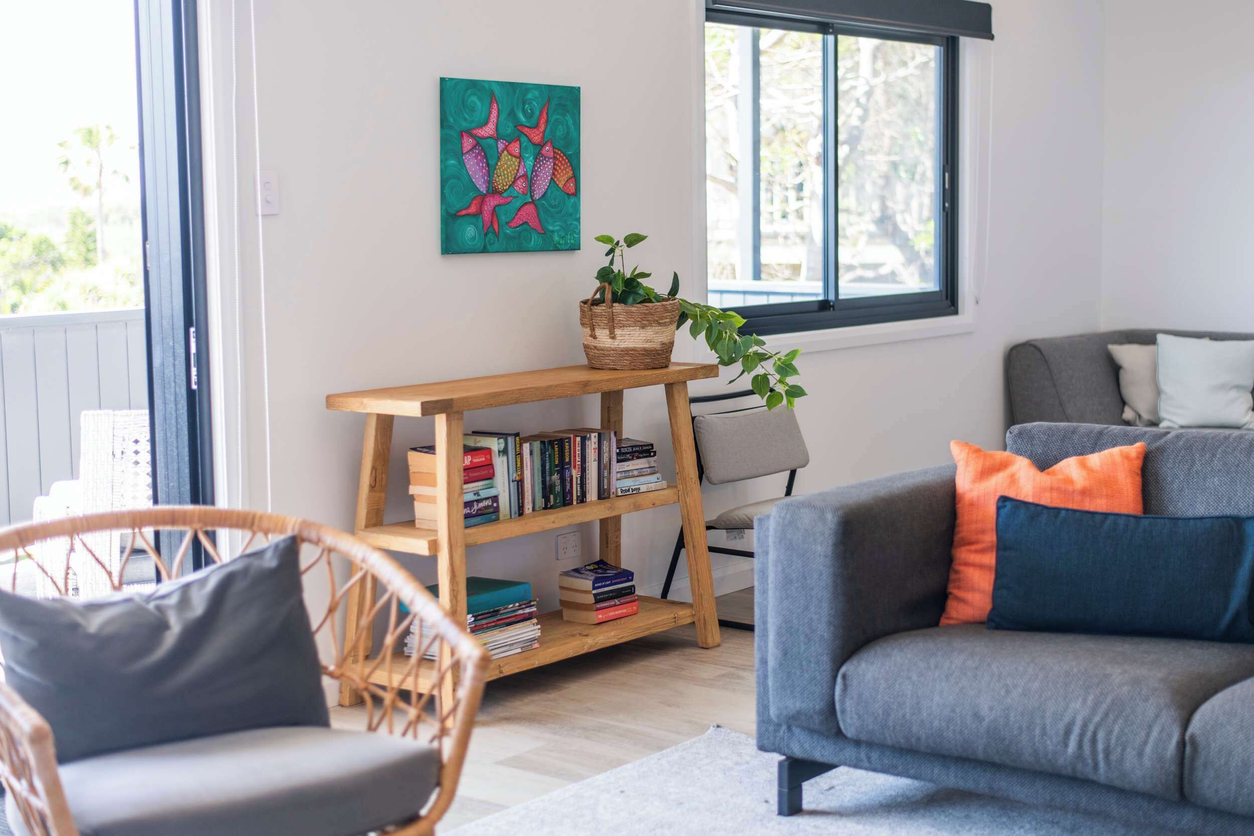 Una sala de estar con un sofá gris, una silla de mimbre, una estantería y una ventana con vistas.