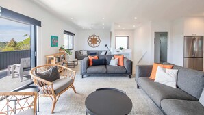 Ein modernes Wohnzimmer mit einem grauen Sofa, einem Korbstuhl, einem schwarzen Couchtisch und einem grauen Teppich. Es gibt eine Küchenzeile mit einem Kühlschrank sowie einen Esstisch mit Stühlen.