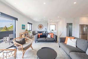 A modern living room with a grey sofa, a wicker chair, a black coffee table, and a grey rug. There is a kitchen area with a refrigerator and a dining table with chairs.