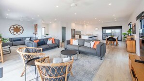 Ein modernes Wohnzimmer mit einer grauen Sofagarnitur, Korbstühlen, einem Couchtisch und einer Küche im Hintergrund.