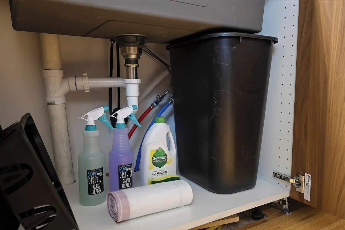 A kitchen sink with various cleaning supplies and a black trash bin.