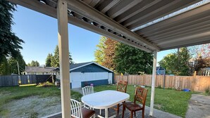 A covered patio with a table and chairs, a wooden deck, and a backyard with a lawn and a wooden fence.