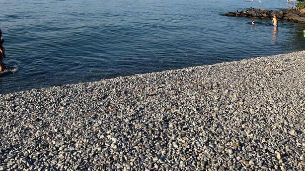 Una spiaggia di ciottoli con acqua calma, colline in lontananza e cielo sereno.