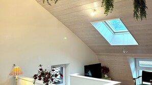 Un interno moderno con soffitto alto, travi in legno, un lucernario, una reception, una TV e piante sospese.