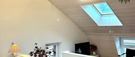 Ein modernes Interieur mit hoher Decke, Holzbalken, einer Dachfensterkuppel, einem Empfangstresen, einem Fernseher und hängenden Pflanzen.