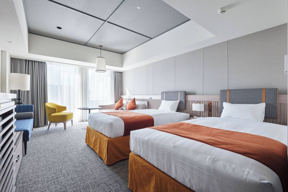 A hotel room with two beds, a desk, a chair, and a large window with curtains.