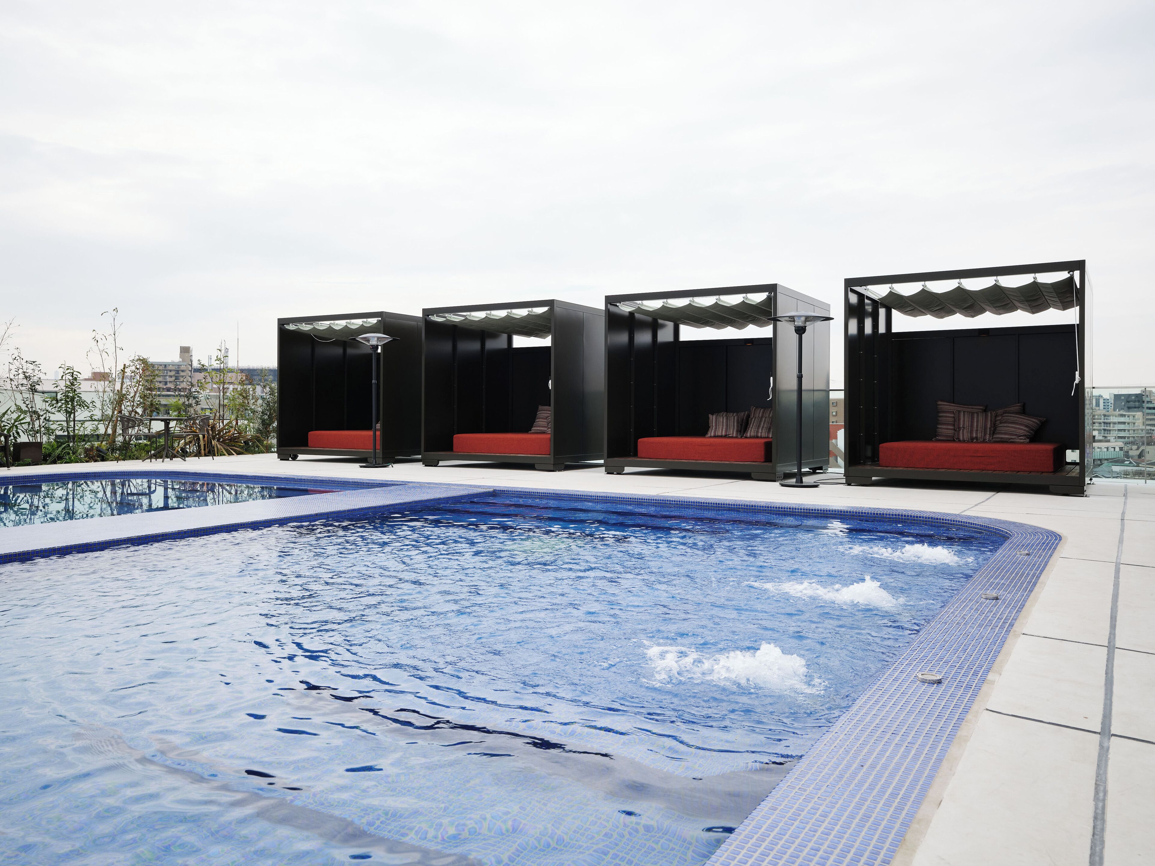 Un espace piscine aménagé sur le toit, comprenant quatre cabanas noires, chacune dotée d’un coussin rouge et d’un oreiller.
