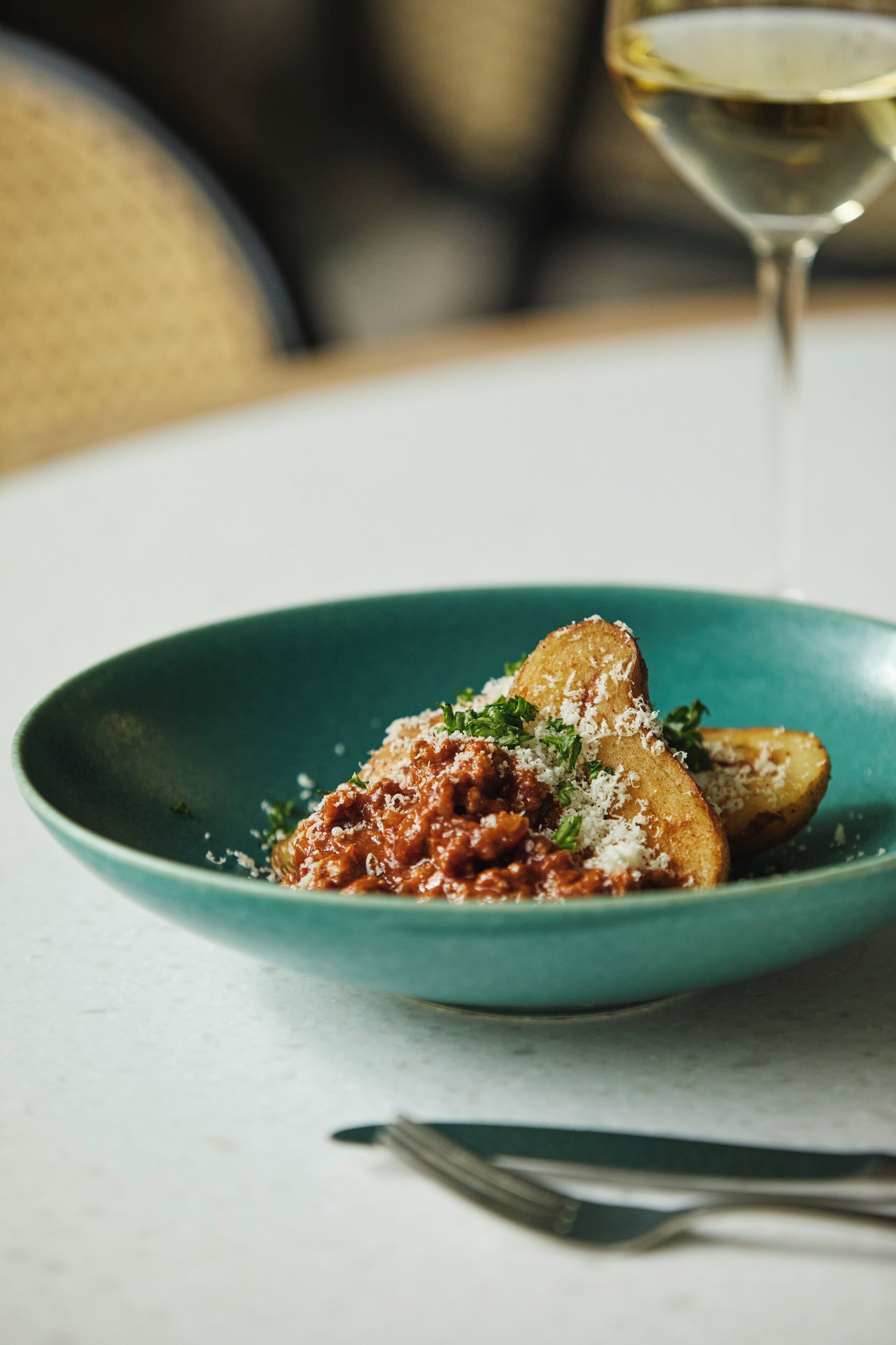 A teal bowl with a pasta dish, a fork, and a glass of white wine on a table.