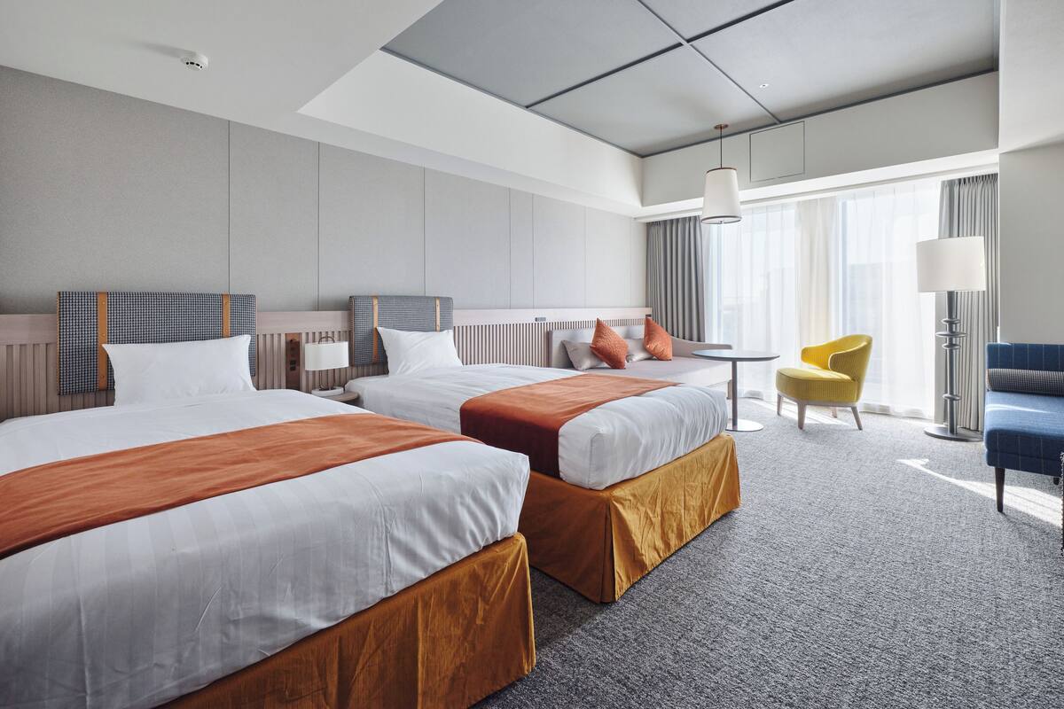A modern hotel room with two beds, a sofa, a chair, and a lamp.