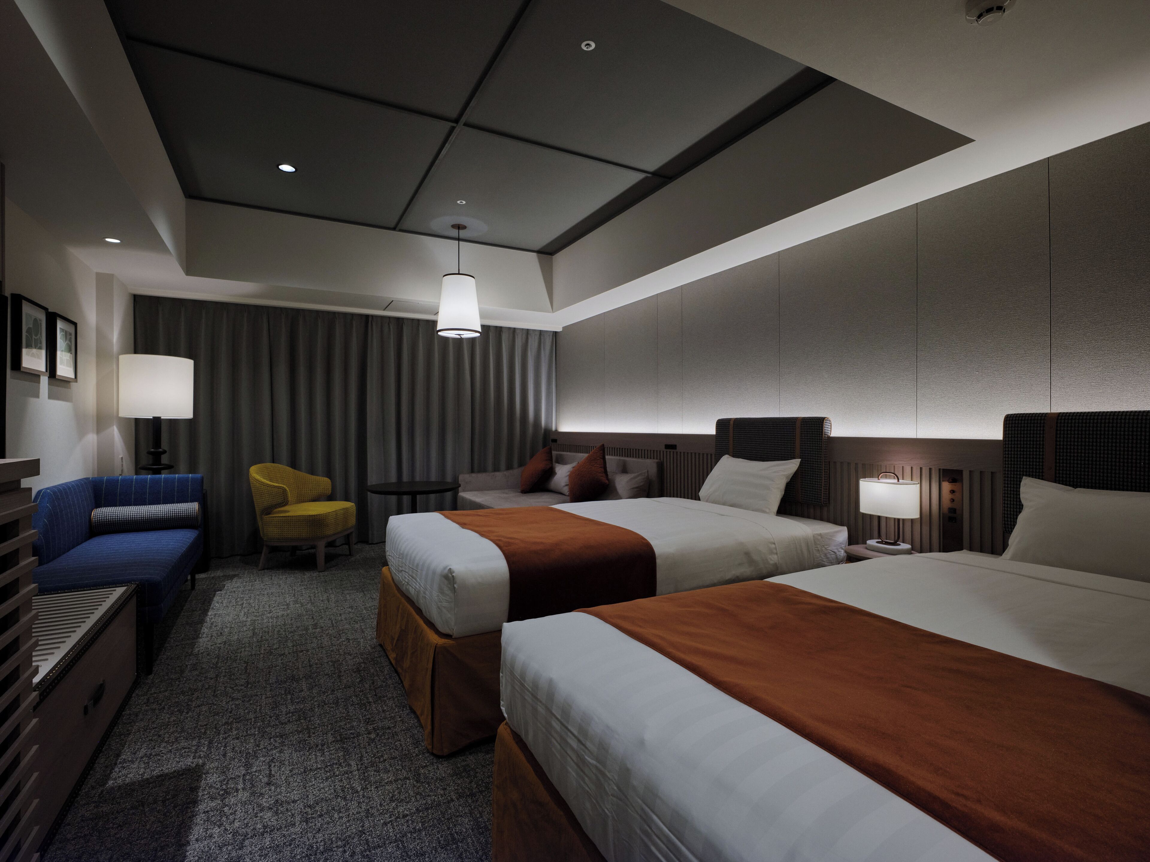 A modern hotel room with two beds, a sofa, a chair, and a small table.