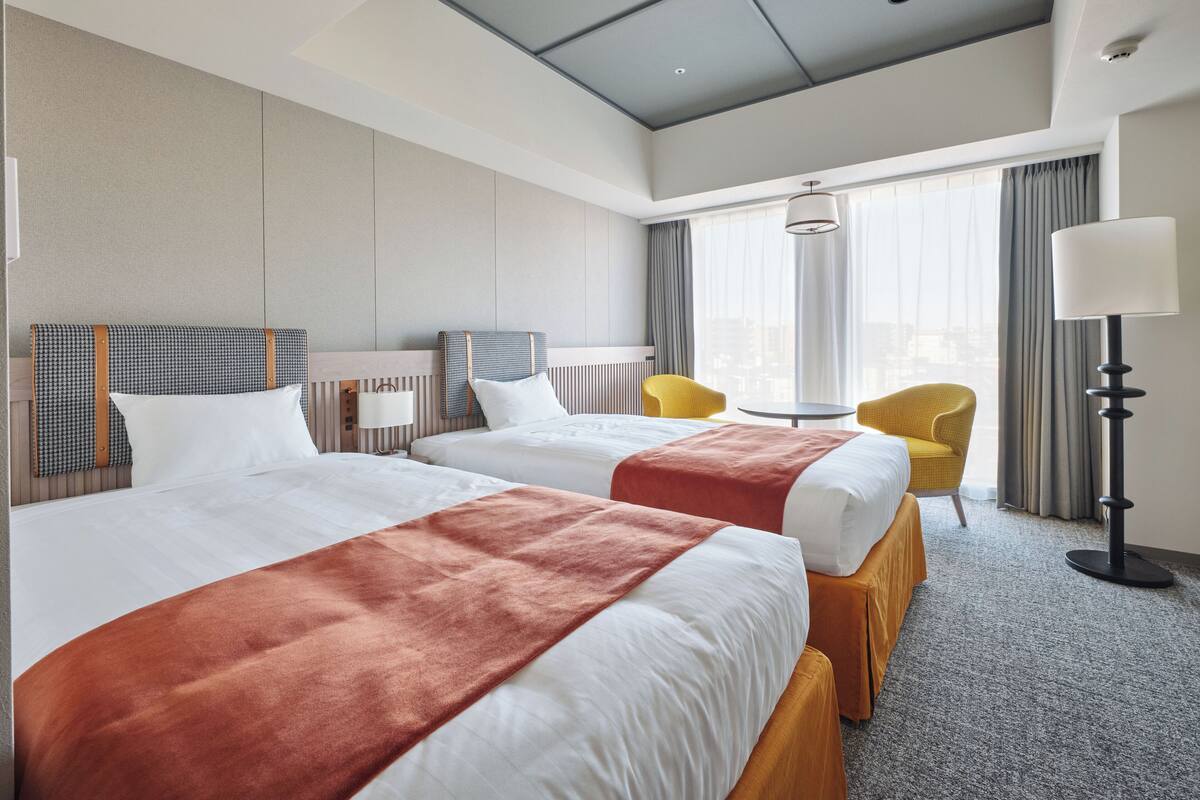 A hotel room with two beds, a small table, a chair, and a lamp.