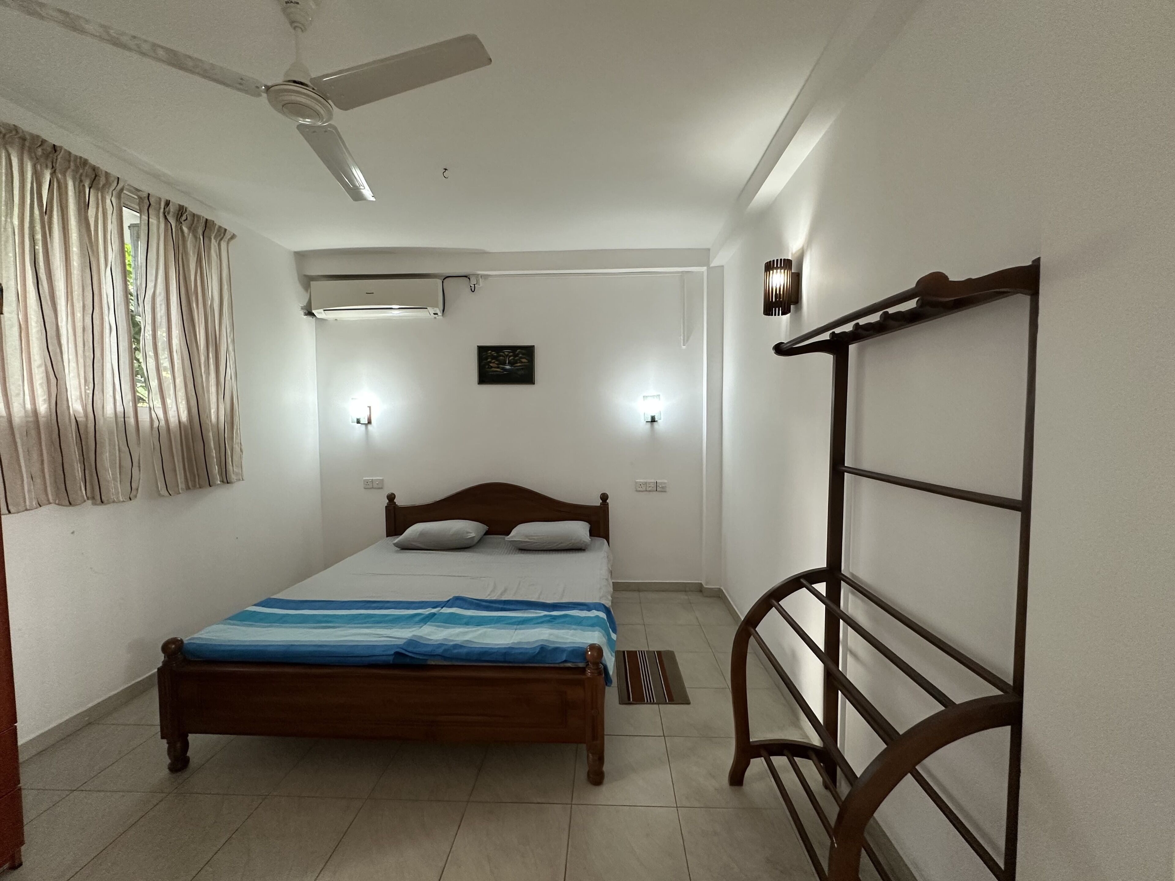 A bedroom with a bed, ceiling fan, air conditioning unit, bedside lamps, and a window with curtains.