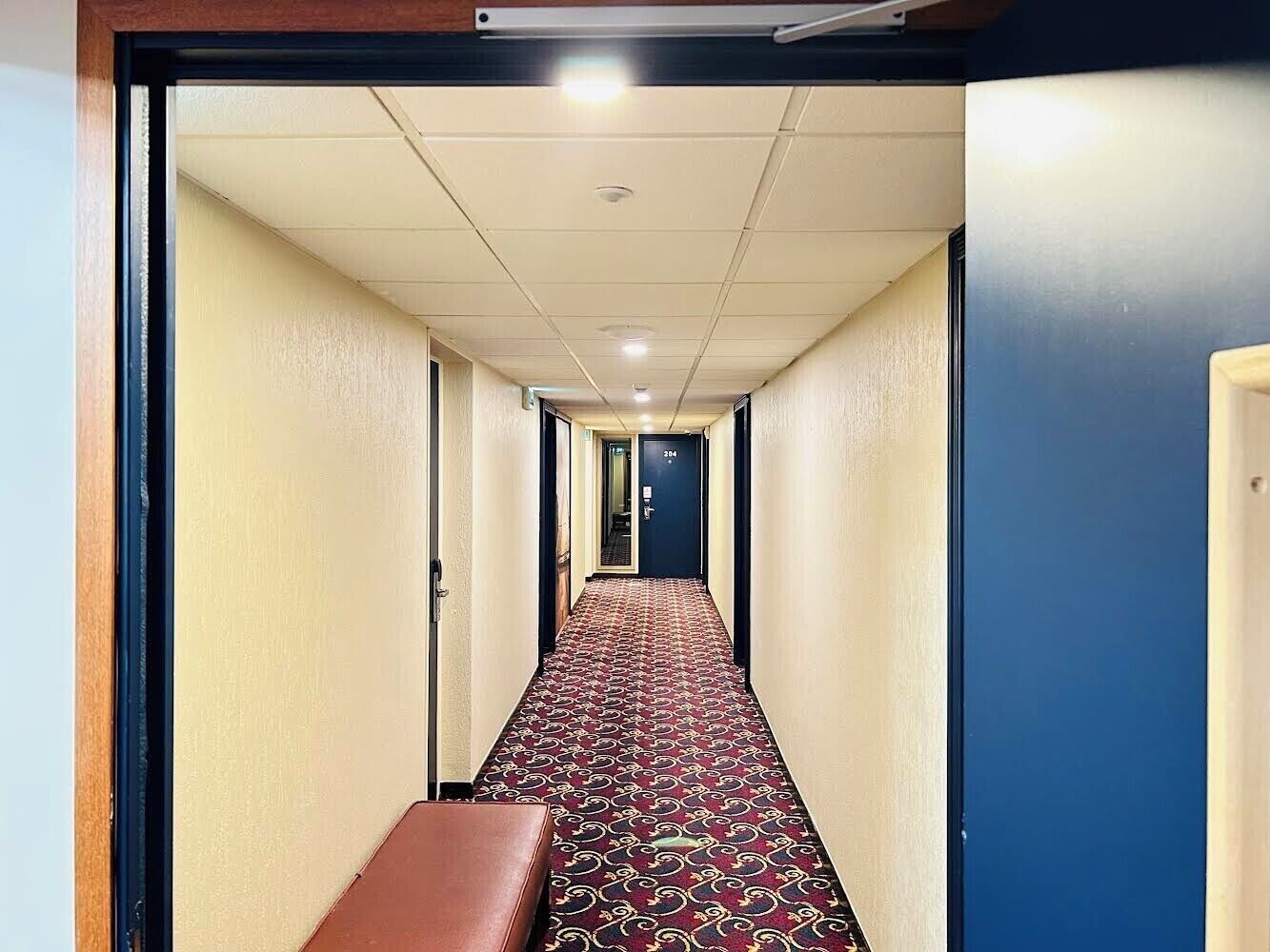 Een hotelgang met een gekleurd tapijt, een bankje en deuren naar kamers.