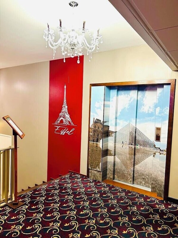 Un vestíbulo de hotel con una pared roja decorativa, un candelabro, una puerta de cristal grande con el reflejo de un edificio y una alfombra estampada.